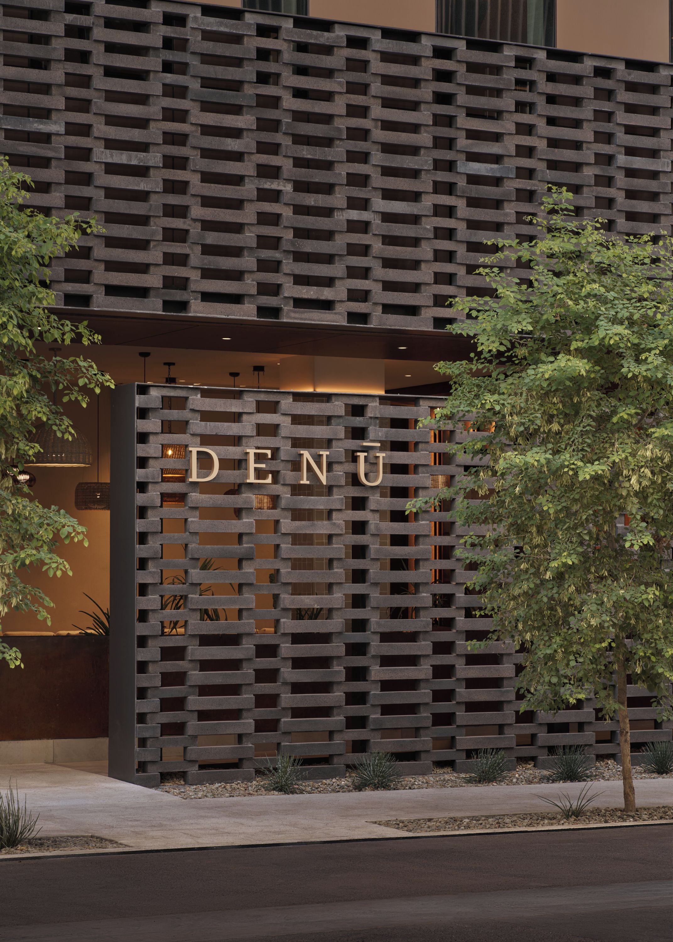 Fachada de edificio con diseño moderno, que presenta un patrón similar a una cuadrícula de bloques de concreto y una entrada destacada con la palabra 'DENU' iluminada.