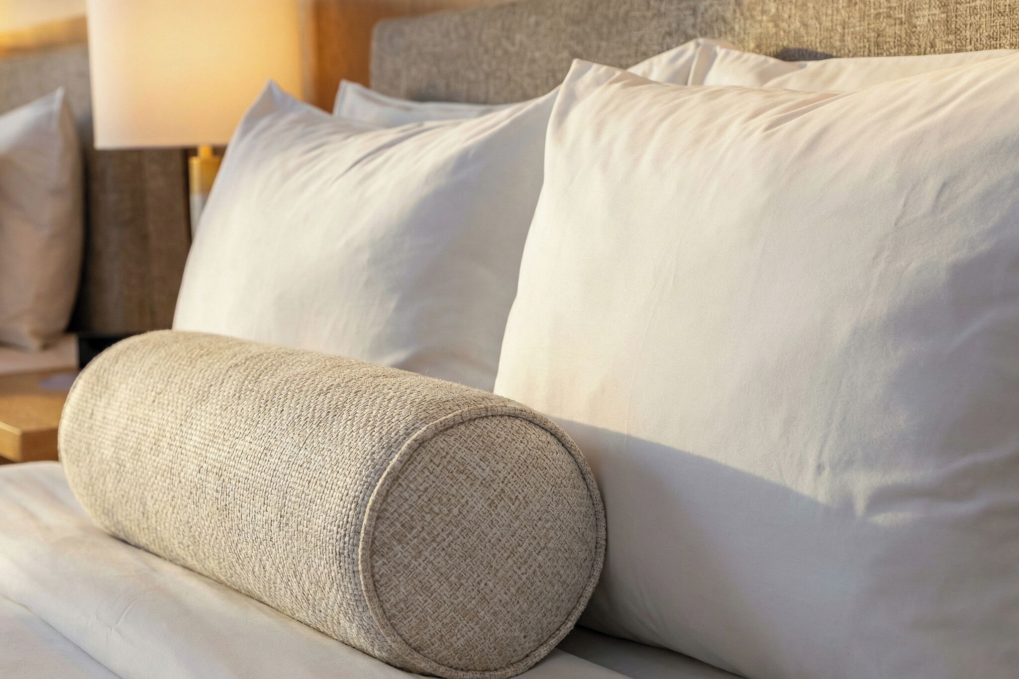 A bed with white linens, a rolled-up beige textured pillow, and a matching pillow on a headboard.