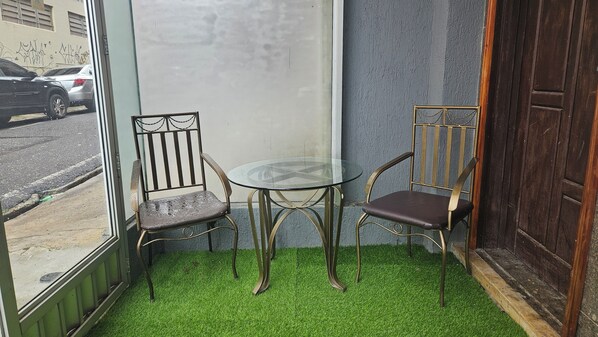 Un petit coin salon avec deux chaises et une table en verre, entouré d’un sol recouvert d’un gazon artificiel vert, d’une fenêtre et d’une porte.
