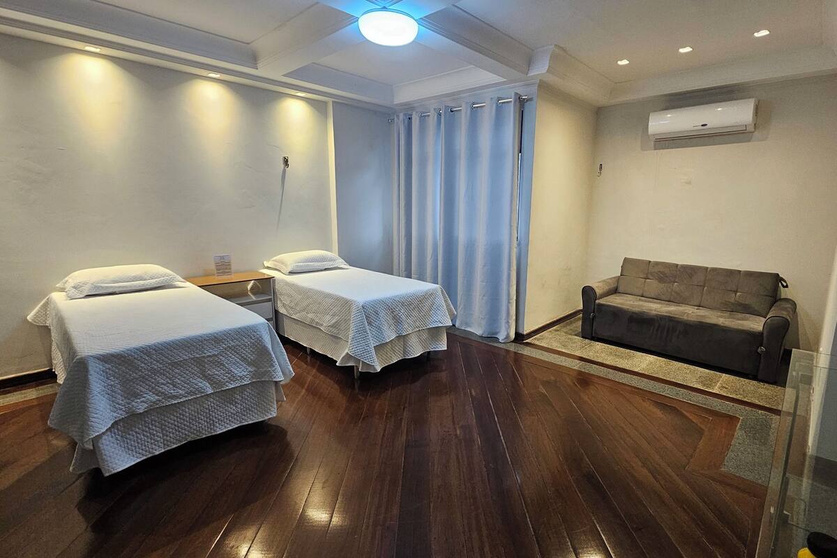 A hotel room with two single beds, a sofa, a small table, and an air conditioning unit.