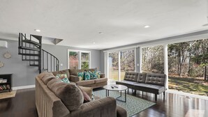 A spacious living room with a curved staircase, large windows, and a view of the outdoors. - Family-Friendly Mashpee Retreat w/ Outdoor Fun (Mashpee)