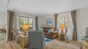 Una habitación bien iluminada con un sofá, un sillón y una pequeña mesa con lámparas. Hay cortinas en las ventanas y un cuadro en la pared.