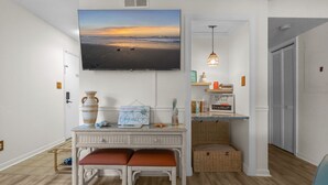 Una habitación con piso de madera, un televisor blanco montado en la pared mostrando una escena de playa, un escritorio de mimbre con un banco y una cesta de mimbre.