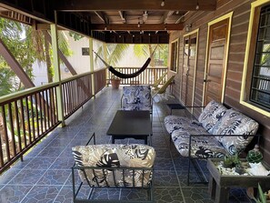 A covered outdoor seating area with a hammock, patterned sofa, and chairs, surrounded by wooden walls and a tiled floor. - Driftwood Gardens Guesthouse: Spacious, fully-equipped 2 bedroom Value Apartment (Placencia)