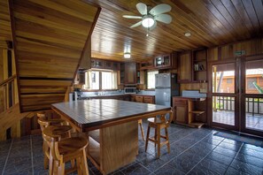 A wooden interior with a kitchen area, a dining table with stools, and a ceiling fan. - Driftwood Gardens Guesthouse: Spacious, fully-equipped 2 bedroom Value Apartment (Placencia)