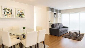 Una sala moderna con una mesa de comedor de vidrio, sillas blancas, un sofá gris y una pequeña mesa de centro. Hay dos cuadros abstractos en la pared y estanterías empotradas.
