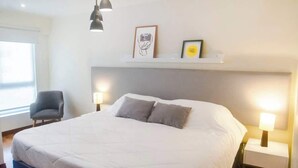Una cama bien hecha con dos almohadas, una lámpara de noche y una silla en una habitación luminosa, con cabecera blanca y estante sobre la cama.
