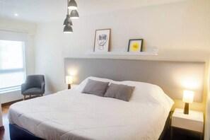A neatly made bed with two pillows, a bedside lamp, and a chair in a well-lit room with a white headboard and a shelf above the bed. - 2 APTS in One Building · Ideal for Large Groups! (Miraflores)
