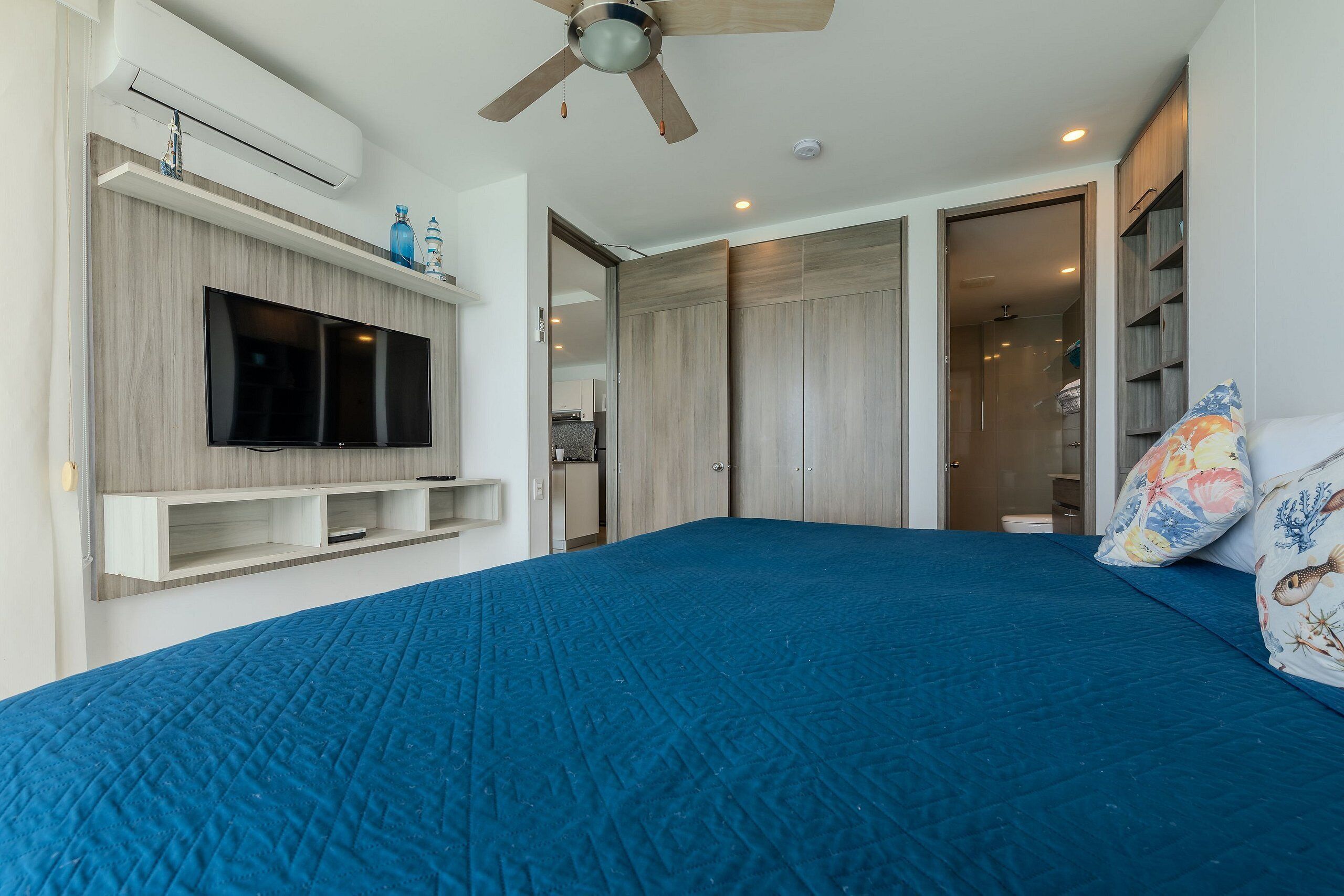 A bedroom with a large bed, a ceiling fan, a TV mounted on a wooden shelf, and a wardrobe with sliding doors.