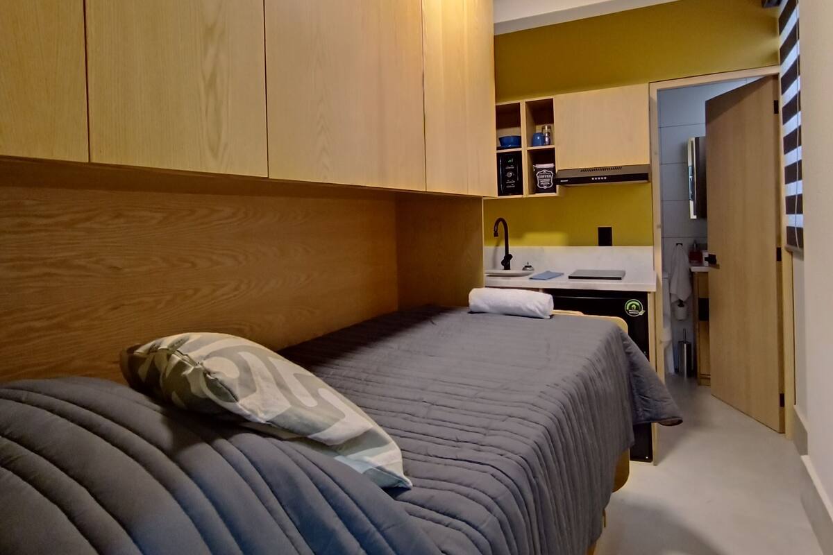 A bedroom with two beds, a wooden wardrobe, a small kitchenette, and a bathroom visible through an open door.