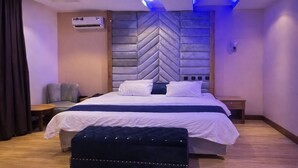 Un dormitorio moderno con una cama grande, un cabecero con estampado en zigzag, un sillón gris, una mesita de noche de madera y una pequeña mesa.