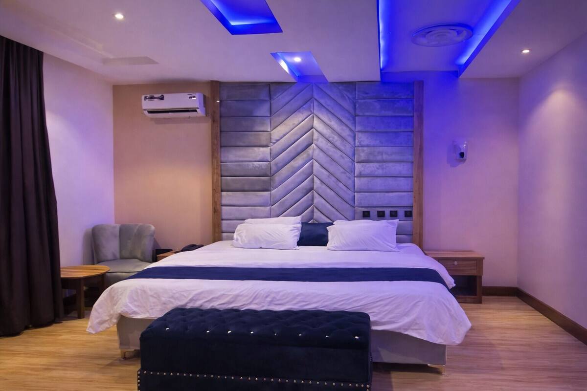Un dormitorio moderno con una cama grande, un cabecero con estampado en zigzag, un sillón gris, una mesita de noche de madera y una pequeña mesa.