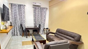 Una sala de estar con un sofá de cuero marrón, una mesita y un televisor sobre un mueble bajo. Se observa una ventana con cortinas y un equipo de aire acondicionado en la pared.