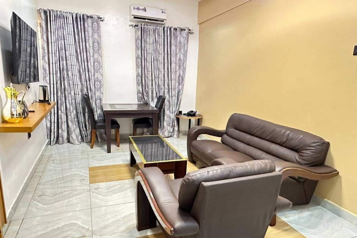 Una sala de estar con un sofá de cuero marrón, una mesita y un televisor sobre un mueble bajo. Se observa una ventana con cortinas y un equipo de aire acondicionado en la pared.