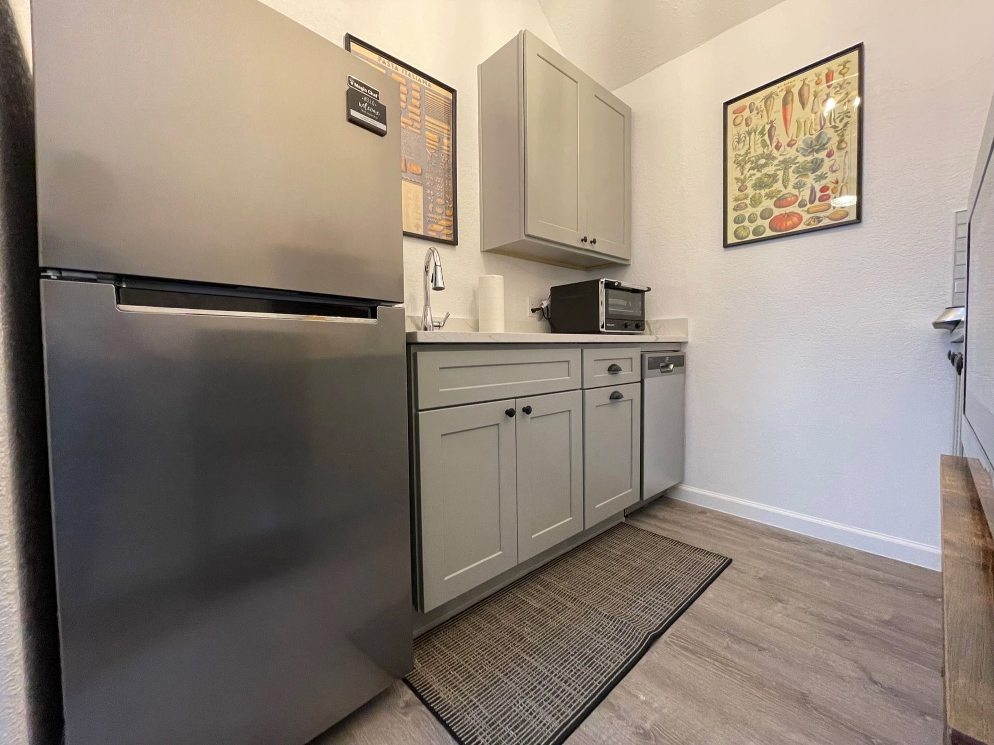 A compact kitchen with a stainless steel refrigerator, a microwave, and a sink. There is a small countertop with a toaster and a cabinet above it. A welcome sign is visible on the wall.