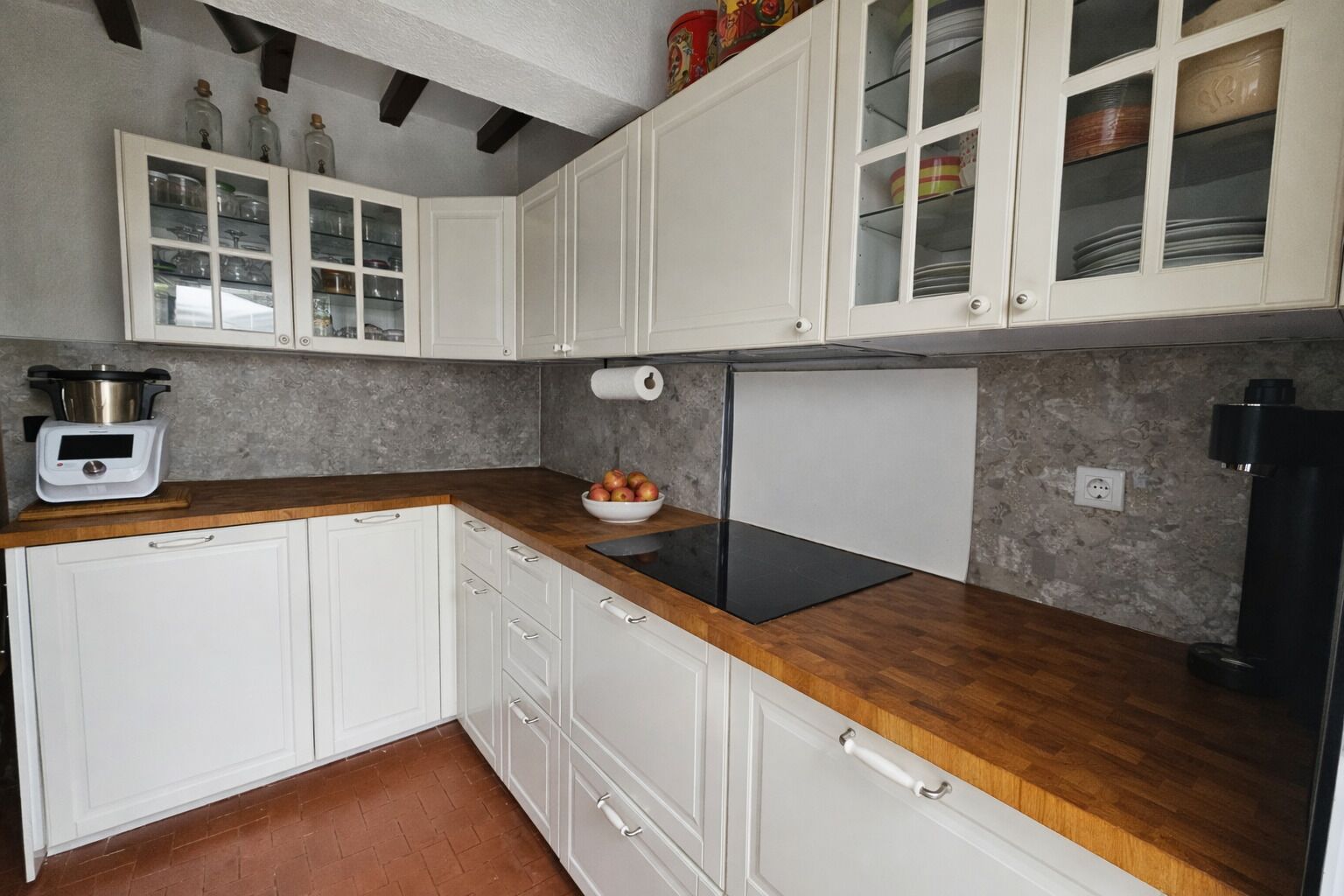 Une cuisine avec des meubles blancs, un plan de travail en bois, une plaque de cuisson à induction noire et un saladier rempli de fruits.