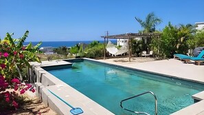 En swimmingpool med utsikt över havet, omgiven av tropiska växter och en liten lusthus.