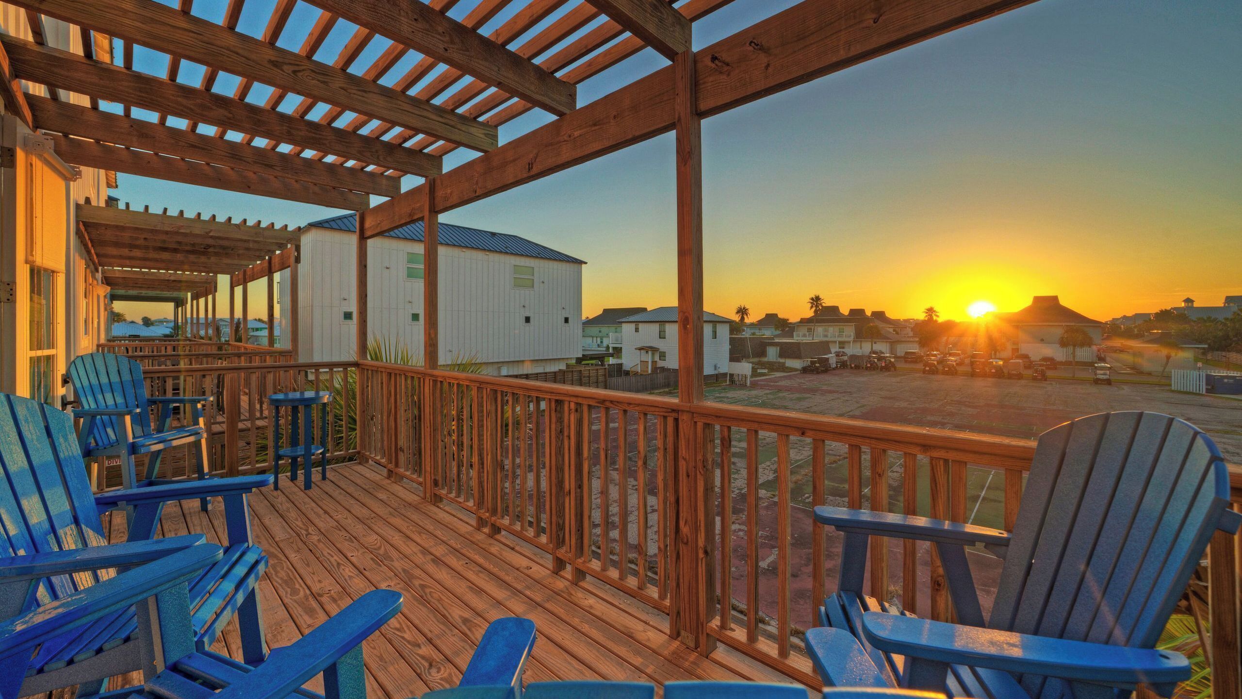 Eine Holzterrasse mit blauen Adirondack-Stühlen, einer Pergola und Blick auf Häuser und den Sonnenuntergang.