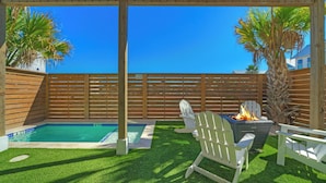 A backyard with a pool, two Adirondack chairs, a fire pit, and a wooden fence. - Southern Cross by Avantstay Steps to the Beach, Pool, Fire Pit & Direct Boardwalk Access (Port Aransas)