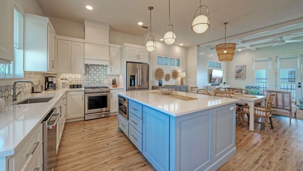 A modern kitchen with white cabinets, stainless steel appliances, a large island, and a dining area with wicker furniture. - Southern Cross by Avantstay Steps to the Beach, Pool, Fire Pit & Direct Boardwalk Access (Port Aransas)