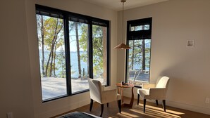 A bedroom with a bed, a desk, two chairs, and large windows overlooking a body of water. - Brand new spa resort style waterfront  executive home with luxury amenities (SALT SPRING ISLAND)