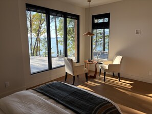 A bedroom with a bed, a desk, two chairs, and large windows overlooking a body of water.