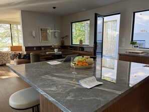 A modern kitchen with a marble countertop, a dining area with a round table, and a large window offering a view of trees.