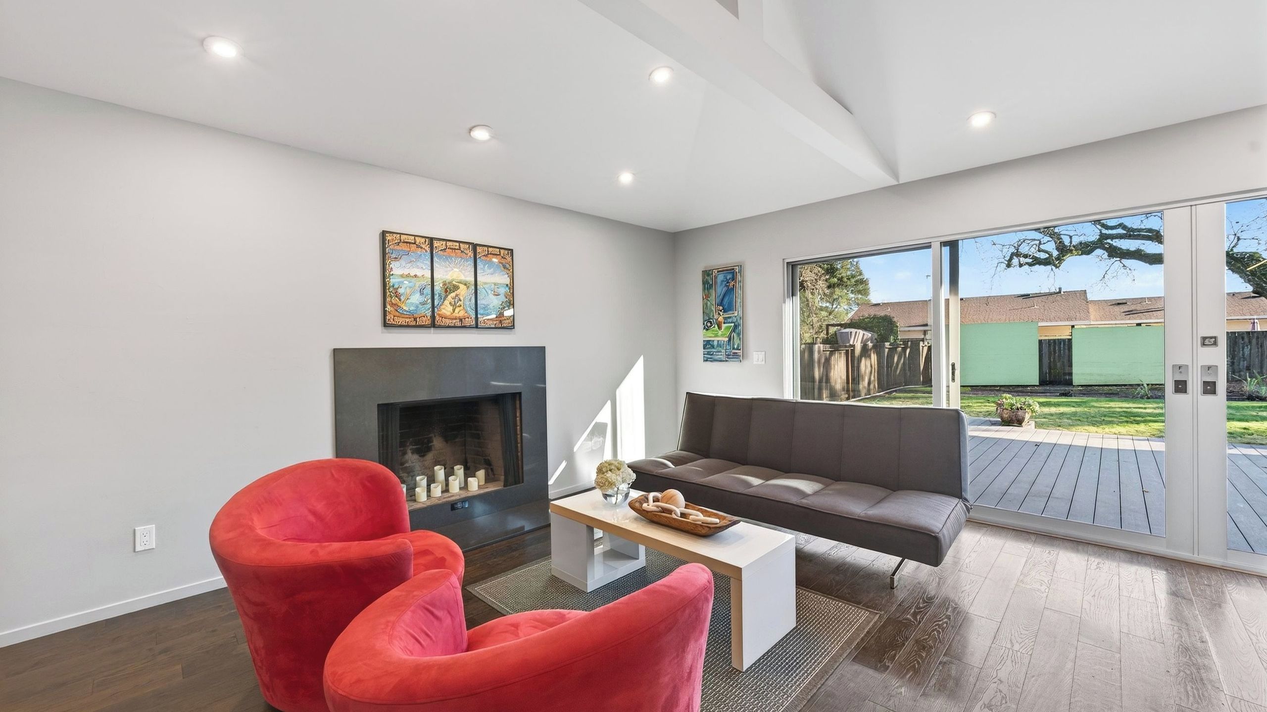 Una sala de estar moderna con chimenea, sillas rojas, un sofá gris y vistas a un edificio verde a través de puertas de cristal amplias.
