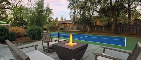 Cancha de tenis exterior con una fogata, rodeada de mobiliario de madera y árboles.