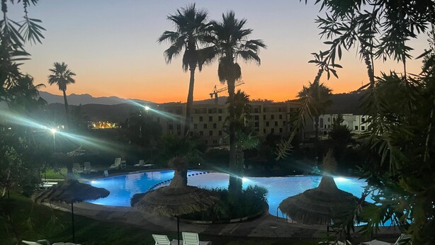 Ein Poolbereich eines Resorts mit zwei Pools, Liegestühlen und Palmen bei Sonnenuntergang.