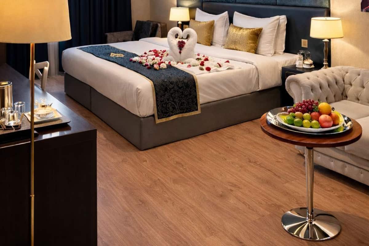 A hotel room with a large bed, a chandelier, a bedside table with a lamp, a round table with fruit, and a dark headboard.