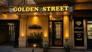 The exterior of the Golden Street hotel with a prominent illuminated sign, a black entrance door, and a small outdoor planter with flowers. - Golden Street Hotel (Baku)