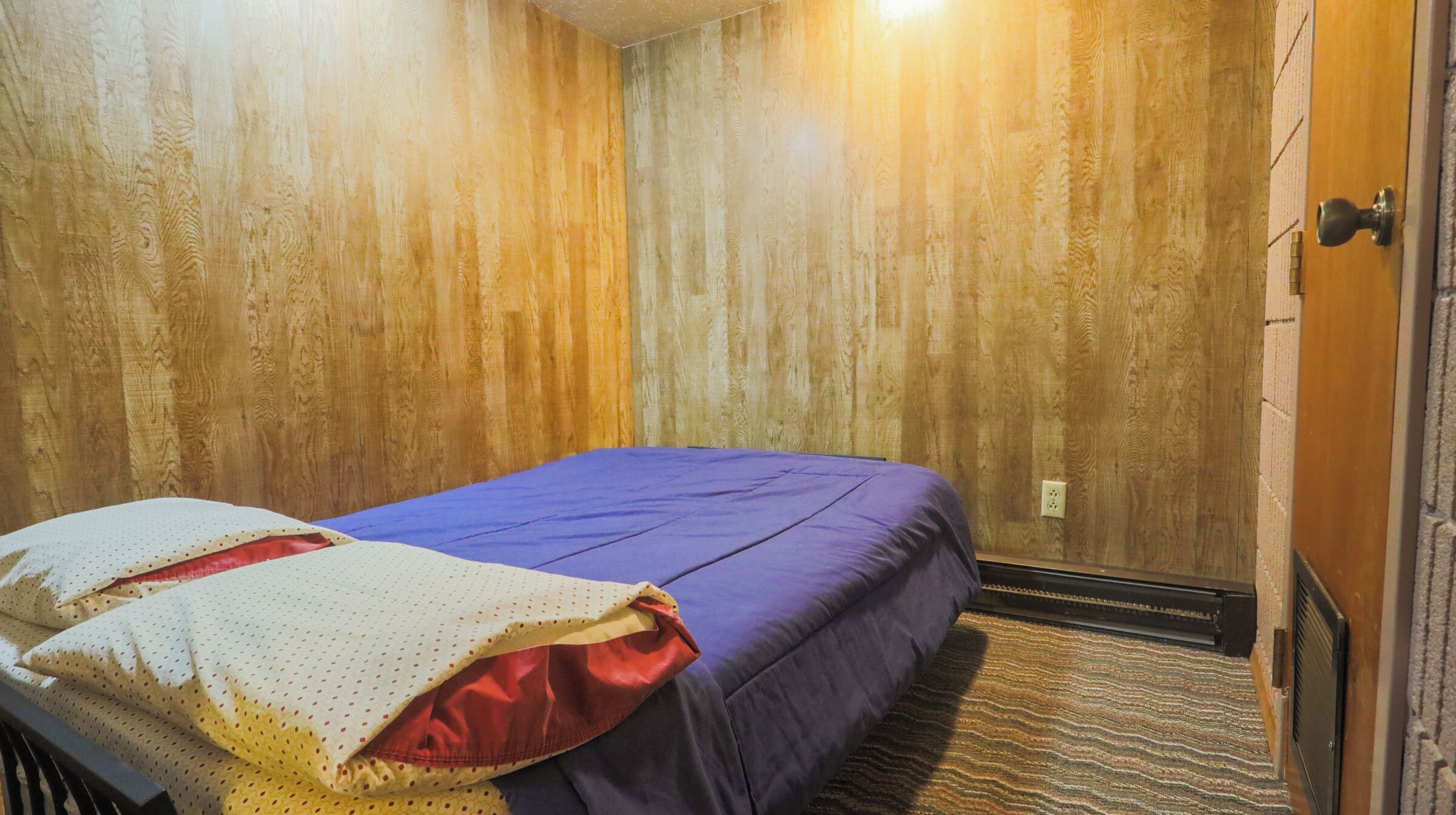 Ein kleines Zimmer mit einem Einzelbett, Holzwänden, einer violetten Bettdecke und einer roten Decke.