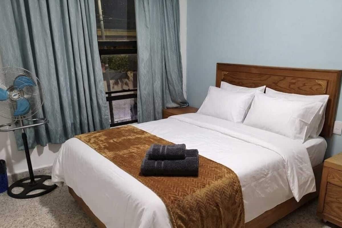 A hotel room with a bed, a wooden headboard, a nightstand, a fan, and a window with curtains.
