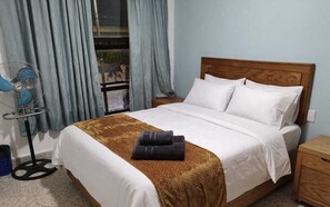 A hotel room with a bed, a wooden headboard, a nightstand, a fan, and a window with curtains. - Mi Casa Elena (Morelia)