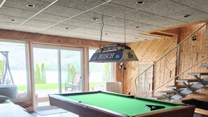 A pool table with a hanging sign, wooden walls, and a view through large windows.