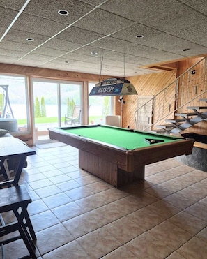 A pool table with a hanging sign, wooden walls, and a view through large windows.