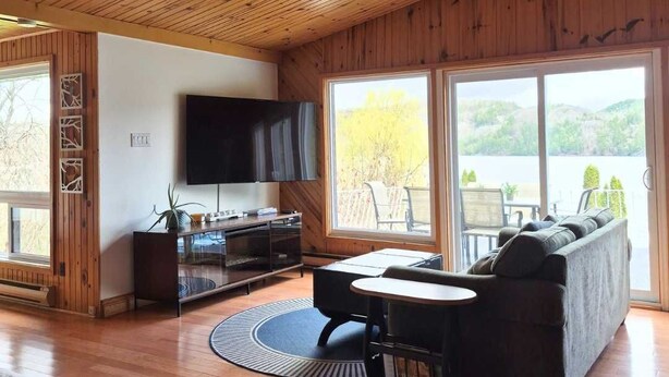A spacious living room with a wooden ceiling, a large TV, a sofa, a coffee table, and a view of a natural landscape through large windows.