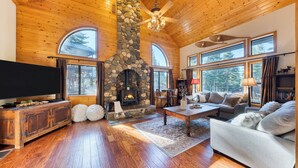 Un salón acogedor con chimenea de piedra, pisos de madera y amplias ventanas que ofrecen vistas al exterior.