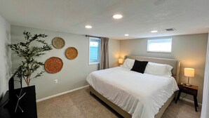 A bedroom with a large bed, a nightstand with a lamp, a window with curtains, and wall decor. - Cozy 2BR ~15 minutes to Red Rocks (Denver)