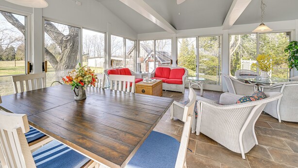 A spacious sunroom with a large wooden table, white wicker furniture, and large windows offering a view of the outdoors. - Bourbon Country Escape! Spacious Home w/ Fire Pit (Taylorsville)