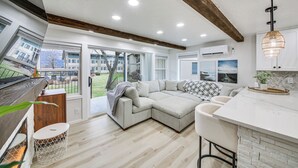 Een moderne woonkamer met een hoekbank, een keuken met een wit aanrecht en uitzicht op de omgeving.