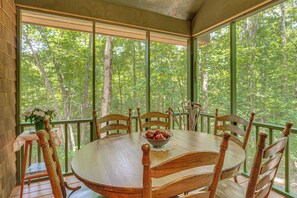 A wooden dining table with chairs, a bowl of fruit, and a view of a forest through large windows. - Vintage Home Nestled in the Woods of Smoky Mtns (Marble)