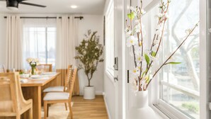 A dining area with a wooden table and chairs, a potted plant, and a window with white curtains. - Hot Tub BBQ Island Outdoor Living Near San Diego Beaches Glidden Glow by Avantstay (San Diego)