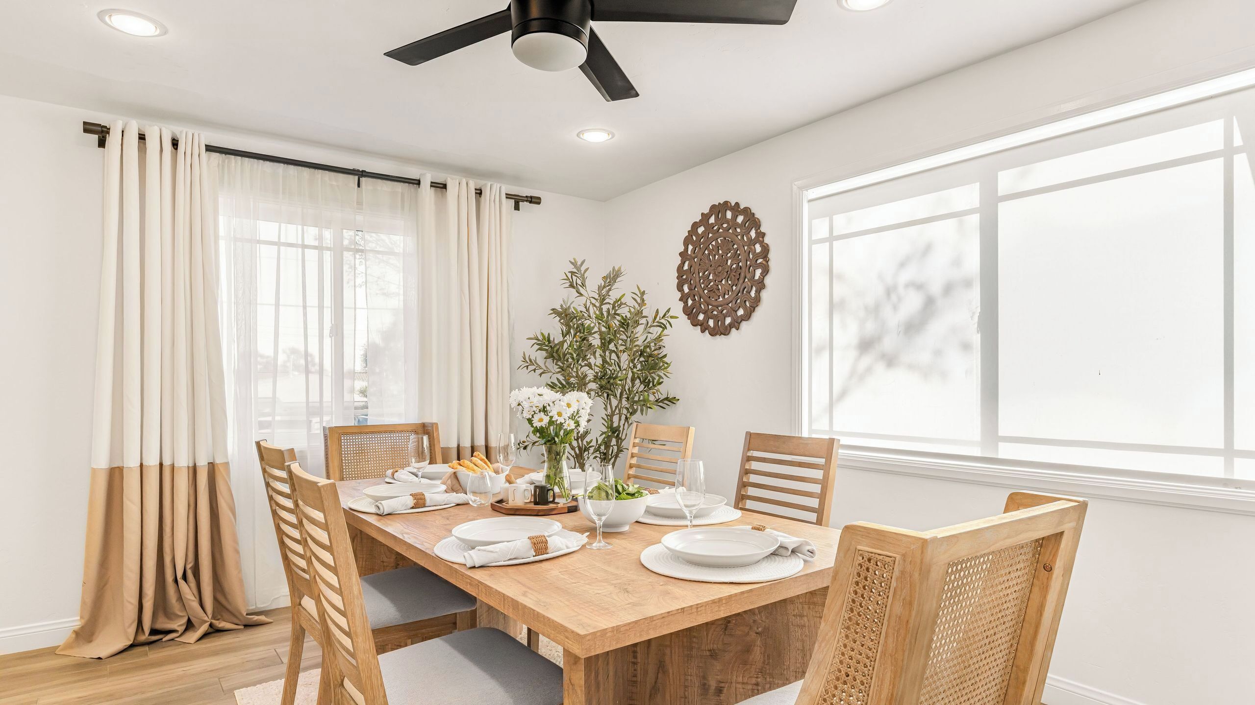A dining room with a wooden table set for four, white chairs, a ceiling fan, and a large window with curtains.
