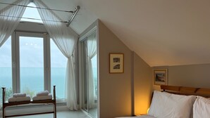 Un dormitorio con una cama grande, un banco de madera con toallas, un ventanal con cortinas translúcidas y un cuadro en la pared.