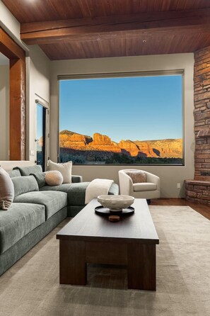 A modern living room with a large window, a sectional sofa, a white armchair, and a wooden coffee table. - 5.5k Sqft Luxury Home with Large Suites, Views, Hot Tub & Firepit (Sedona)
