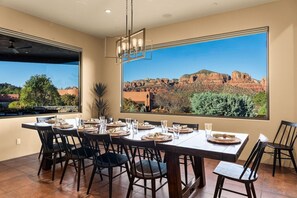 A dining area with a table set for six, large windows with a view of red rock formations, and a modern chandelier. - 5.5k Sqft Luxury Home with Large Suites, Views, Hot Tub & Firepit (Sedona)