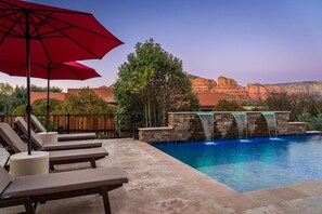 A pool area with lounge chairs, a red umbrella, and a waterfall feature. - 5.5k Sqft Luxury Home with Large Suites, Views, Hot Tub & Firepit (Sedona)