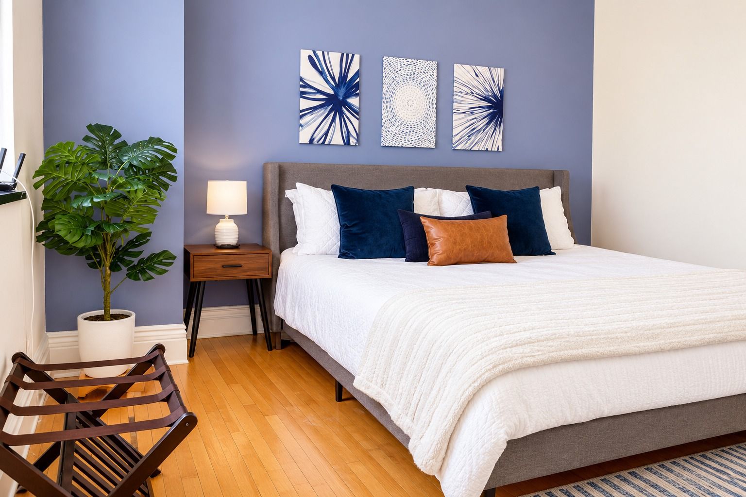 Un dormitorio con cama, mesita de noche, lámpara, planta en maceta y una obra de arte en la pared.